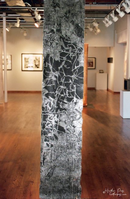 Fantasie by Heather Page is a 60” by 15” monoprint on paper of looping calligraphic text in black and silver on a silver ground that hangs from the ceiling